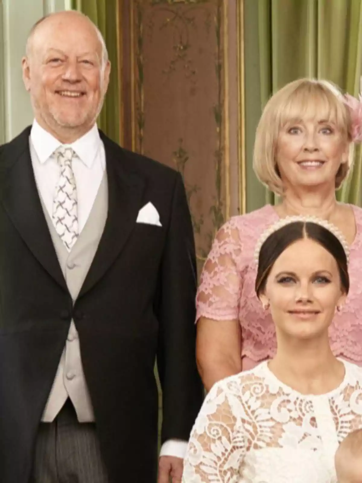 Tres personas posan para una foto formal, dos de pie al fondo y una sentada al frente, todos vestidos elegantemente
