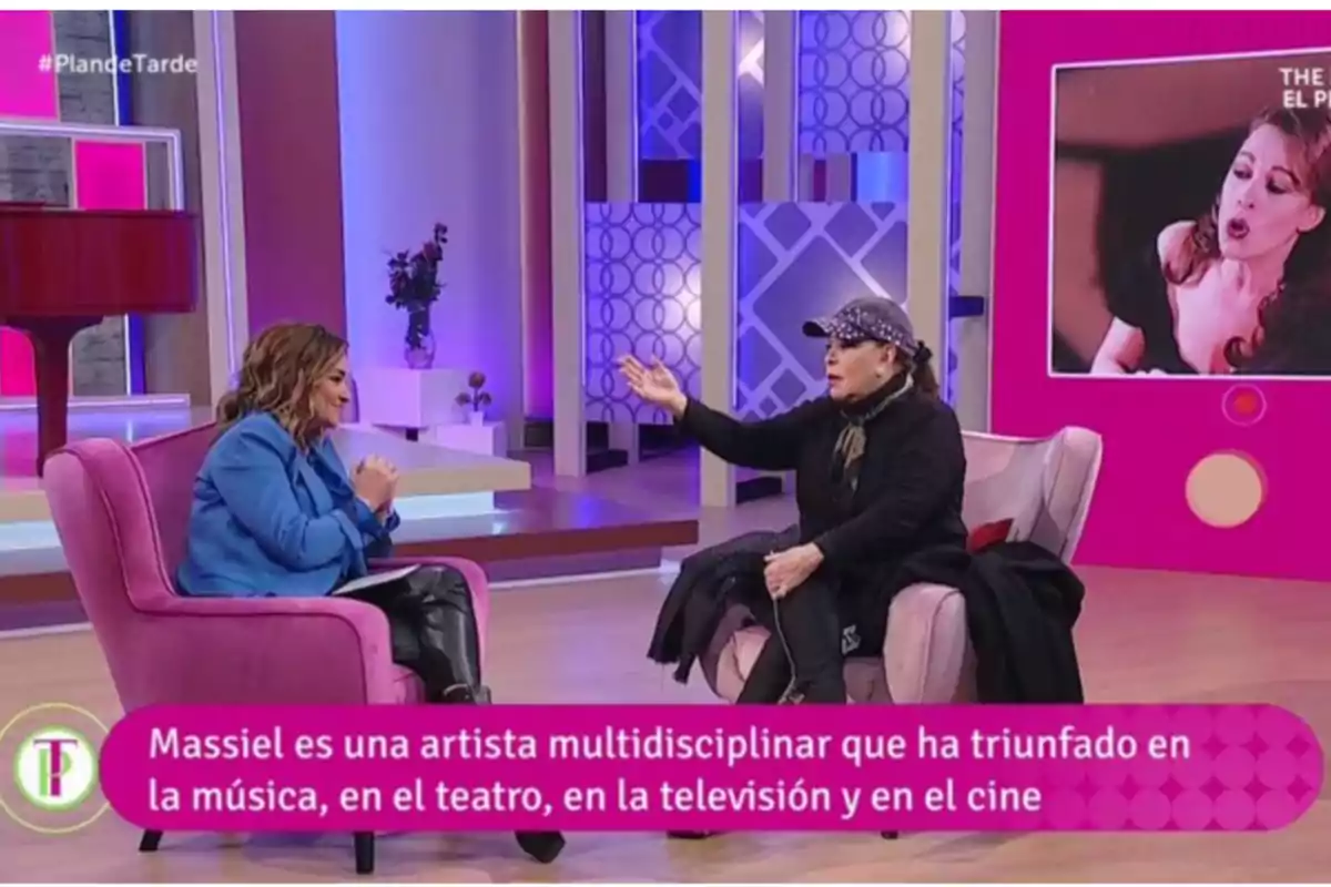 Dos mujeres conversan en un set de televisión decorado en tonos rosas y morados, una de ellas viste chaqueta azul y la otra lleva gorra y ropa oscura, en la pantalla aparece una imagen de una cantante y un texto que destaca la trayectoria multidisciplinar de Massiel en la música, el teatro, la televisión y el cine Dos mujeres conversan en un set de televisión decorado en tonos rosas y morados, una de ellas viste chaqueta azul y la otra lleva gorra y ropa oscura, en la pantalla aparece una imagen de una cantante y un texto que destaca la trayectoria multidisciplinar de Massiel en la música, el teatro, la televisión y el cine