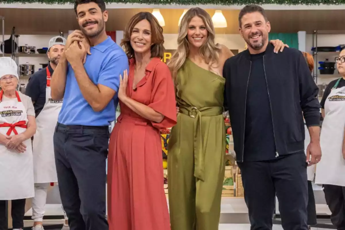 Cuatro personas posan sonrientes al frente de un set de cocina con concursantes y utensilios al fondo Cuatro personas posan sonrientes al frente de un set de cocina con concursantes y utensilios al fondo