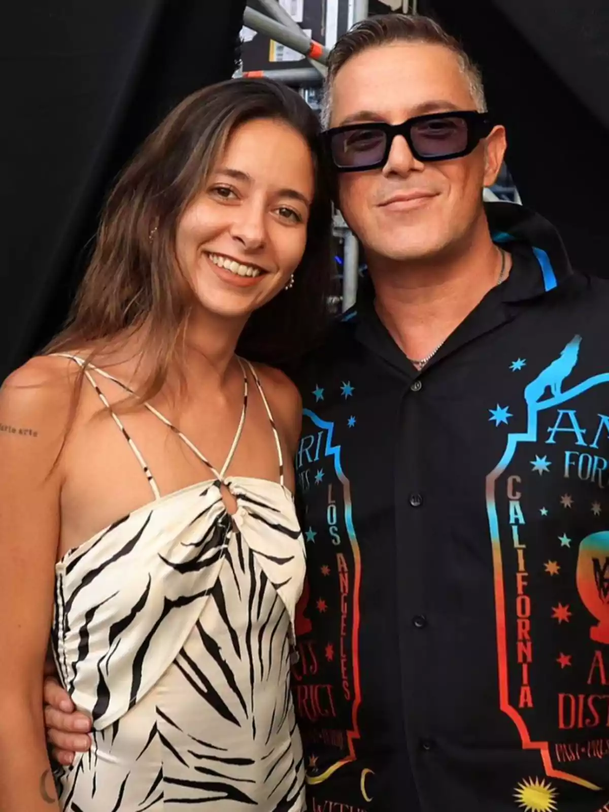 Una mujer con vestido de estampado animal y un hombre con gafas oscuras y camisa negra con letras coloridas posan juntos y sonríen frente a la cámara Una mujer con vestido de estampado animal y un hombre con gafas oscuras y camisa negra con letras coloridas posan juntos y sonríen frente a la cámara