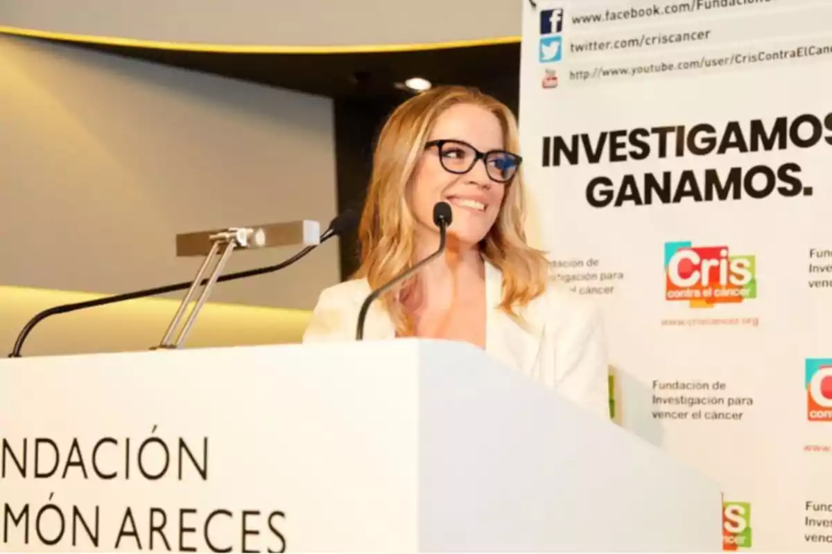 Mujer rubia con gafas sonríe mientras habla en un podio durante un evento de la Fundación Ramón Areces y la organización Cris contra el cáncer con un cartel de fondo que dice investigamos ganamos Mujer rubia con gafas sonríe mientras habla en un podio durante un evento de la Fundación Ramón Areces y la organización Cris contra el cáncer con un cartel de fondo que dice investigamos ganamos