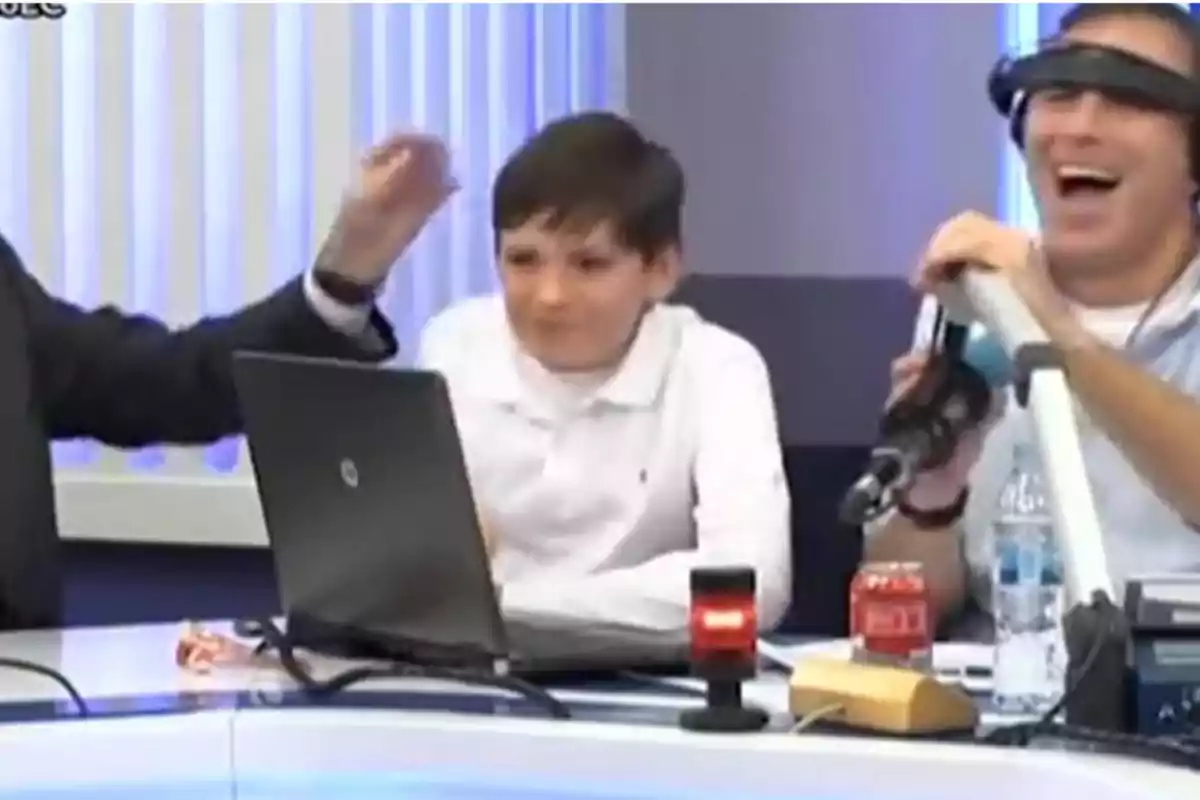 Tres personas en una cabina de radio, una de ellas usando auriculares y riendo, un niño en el centro con expresión seria y una laptop sobre la mesa Tres personas en una cabina de radio, una de ellas usando auriculares y riendo, un niño en el centro con expresión seria y una laptop sobre la mesa