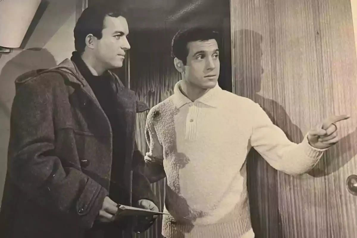 Dos hombres en una escena en blanco y negro, uno de ellos señala hacia la derecha mientras el otro sostiene unos papeles y lo observa. Dos hombres en una escena en blanco y negro, uno de ellos señala hacia la derecha mientras el otro sostiene unos papeles y lo observa.