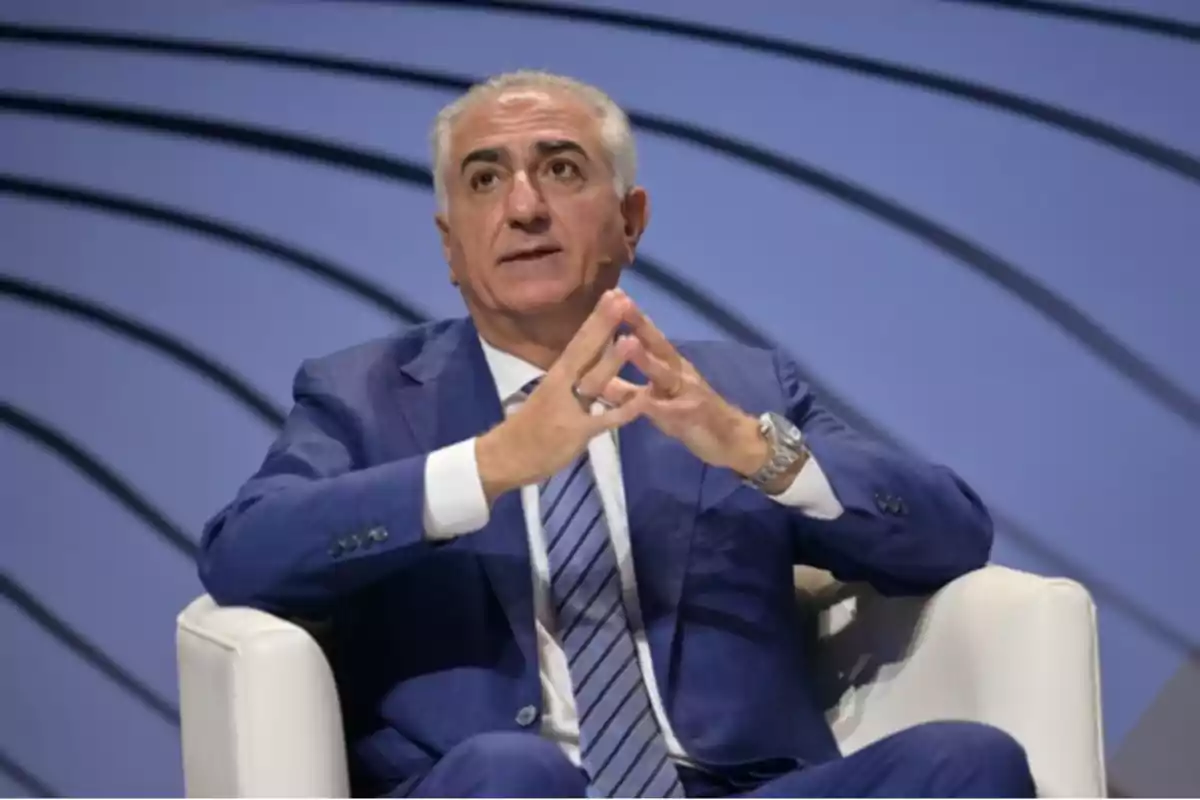 Hombre de cabello canoso con traje azul sentado en un sillón blanco gesticulando con las manos frente a un fondo con líneas curvas