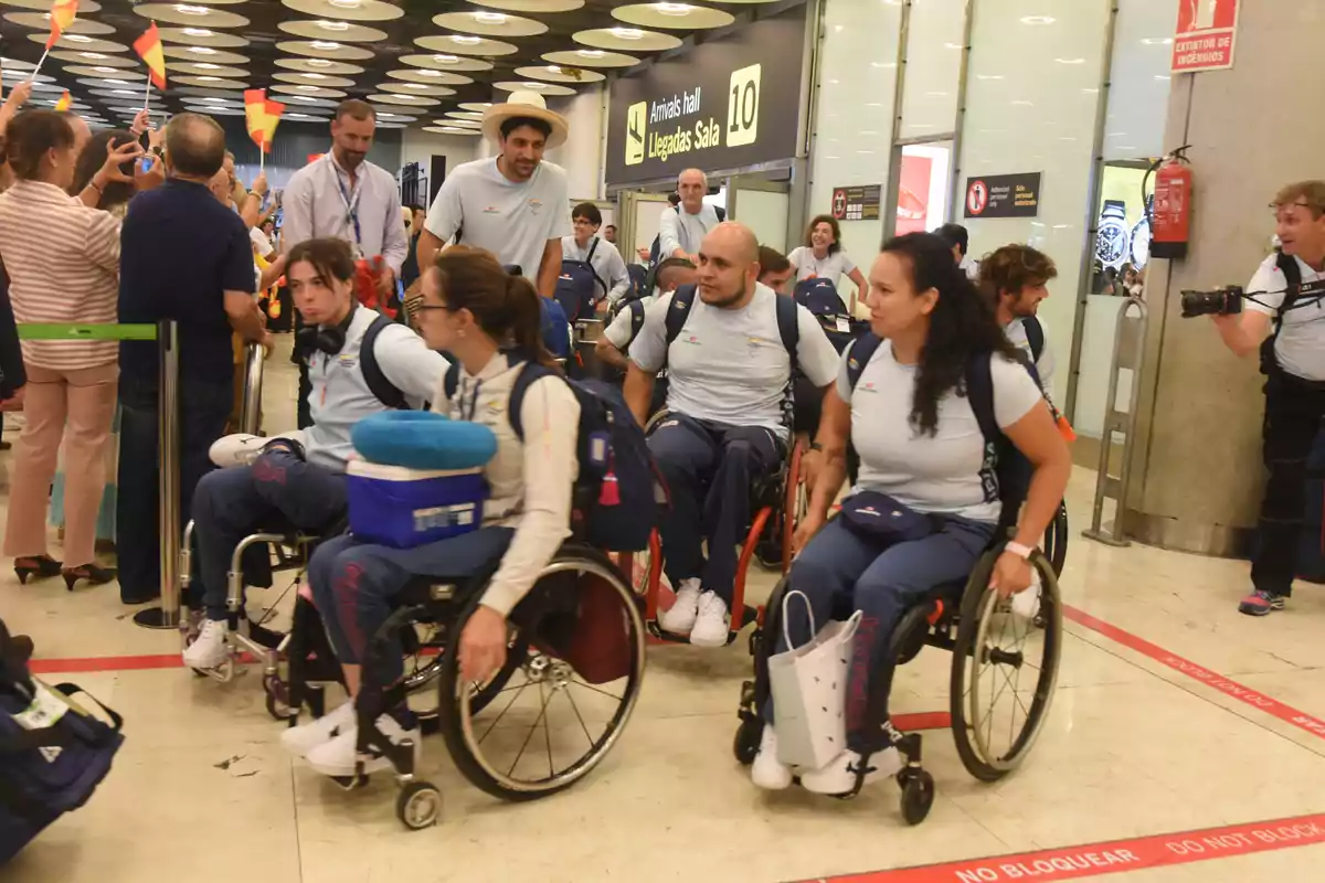 Un grupo de personas en sillas de ruedas, acompañadas por otras personas, se encuentran en una sala de llegadas de un aeropuerto, mientras son recibidas por una multitud que agita banderas.