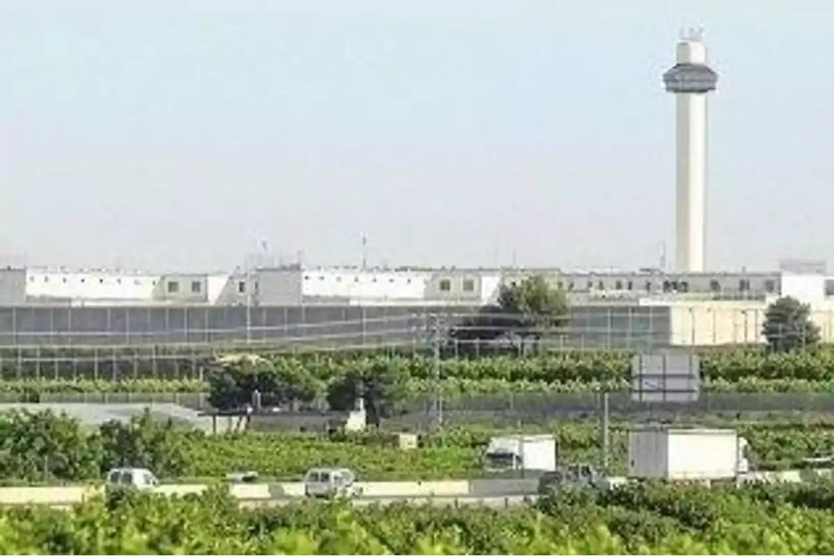 Vista de un edificio grande con una torre alta rodeado de vegetación y vehículos en primer plano. Vista de un edificio grande con una torre alta rodeado de vegetación y vehículos en primer plano.