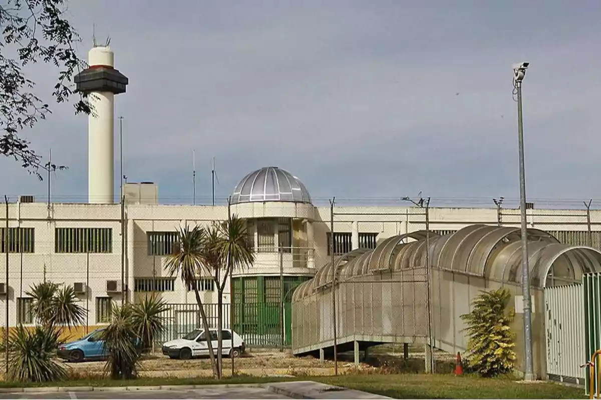 Edificio de una prisión con una torre de vigilancia, cámaras de seguridad y una entrada cubierta por una estructura de metal y vidrio.