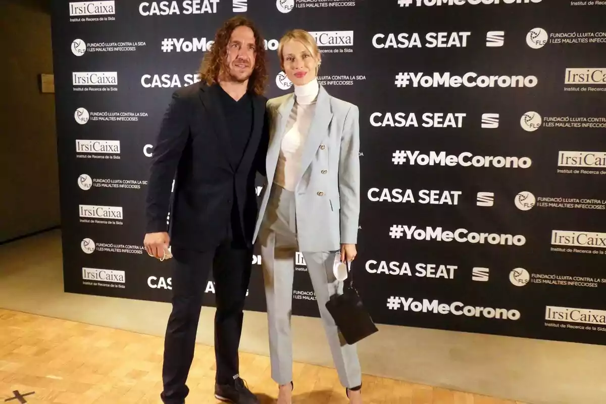 Carles Puyol y Vanesa Lorenzo posan juntos en un evento con un fondo que muestra varios logotipos y hashtags, incluyendo 