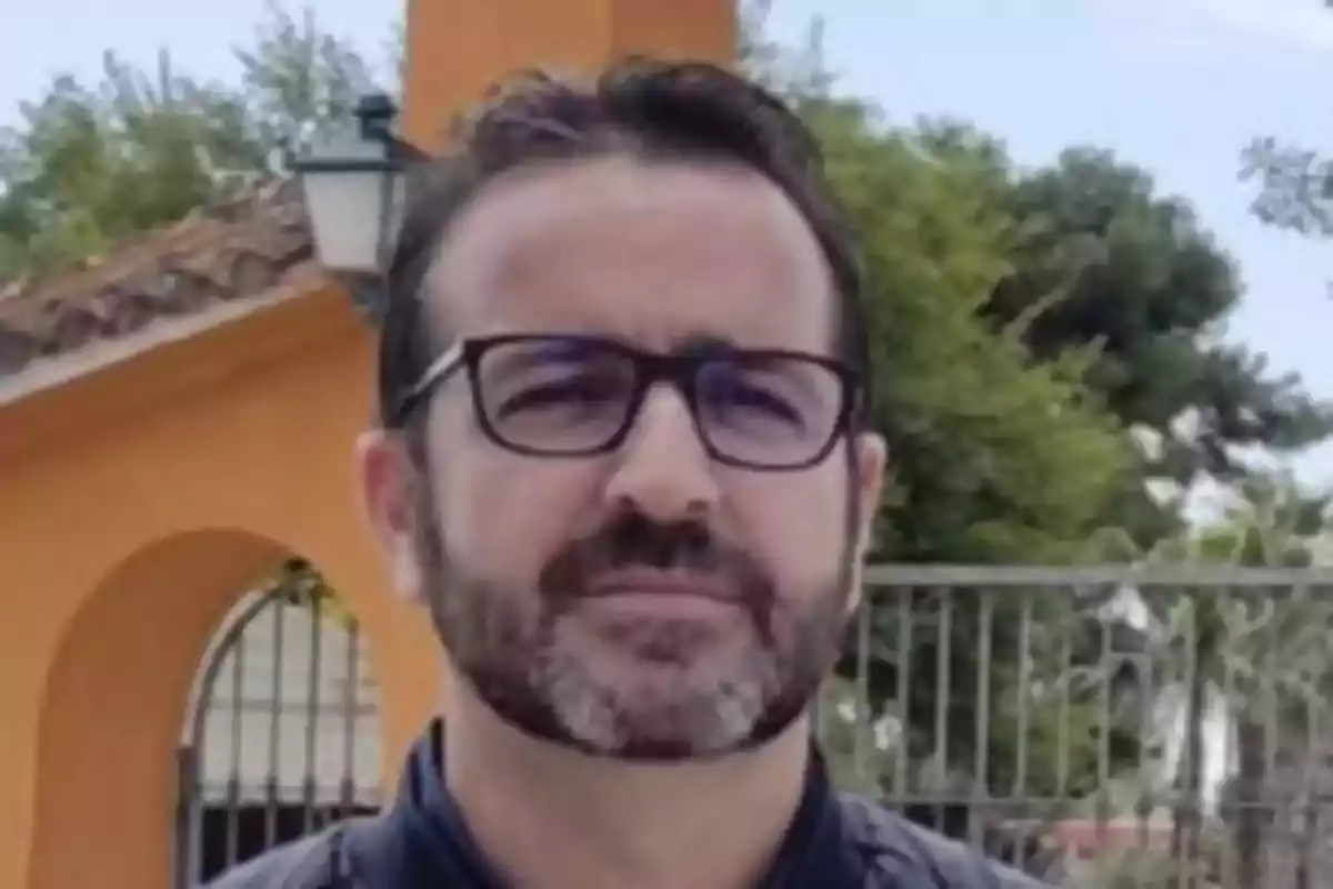 Un hombre con gafas y barba está de pie frente a una estructura de color naranja y un fondo de árboles.