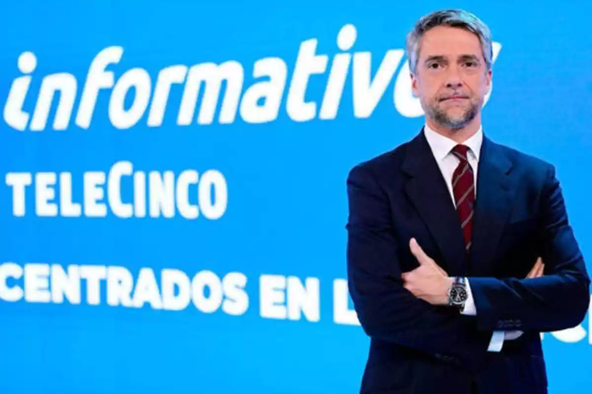 Un hombre de traje y corbata posa con los brazos cruzados frente a un fondo azul que muestra el logotipo de "Informativos Telecinco".
