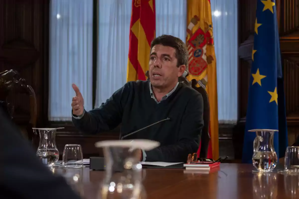 Un hombre hablando en una mesa de reuniones con banderas de fondo.