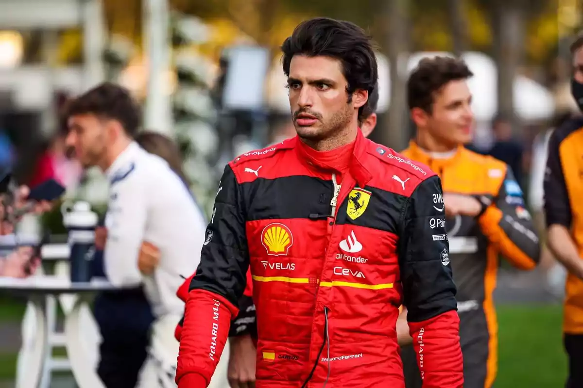 Un piloto de carreras con un traje rojo de Ferrari caminando mientras otras personas están en el fondo.