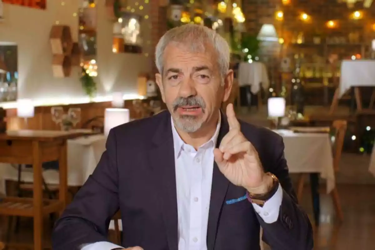 Un hombre de cabello canoso y barba está sentado en un restaurante elegante, vistiendo un traje oscuro y camisa blanca, mientras levanta un dedo como si estuviera haciendo un punto importante.