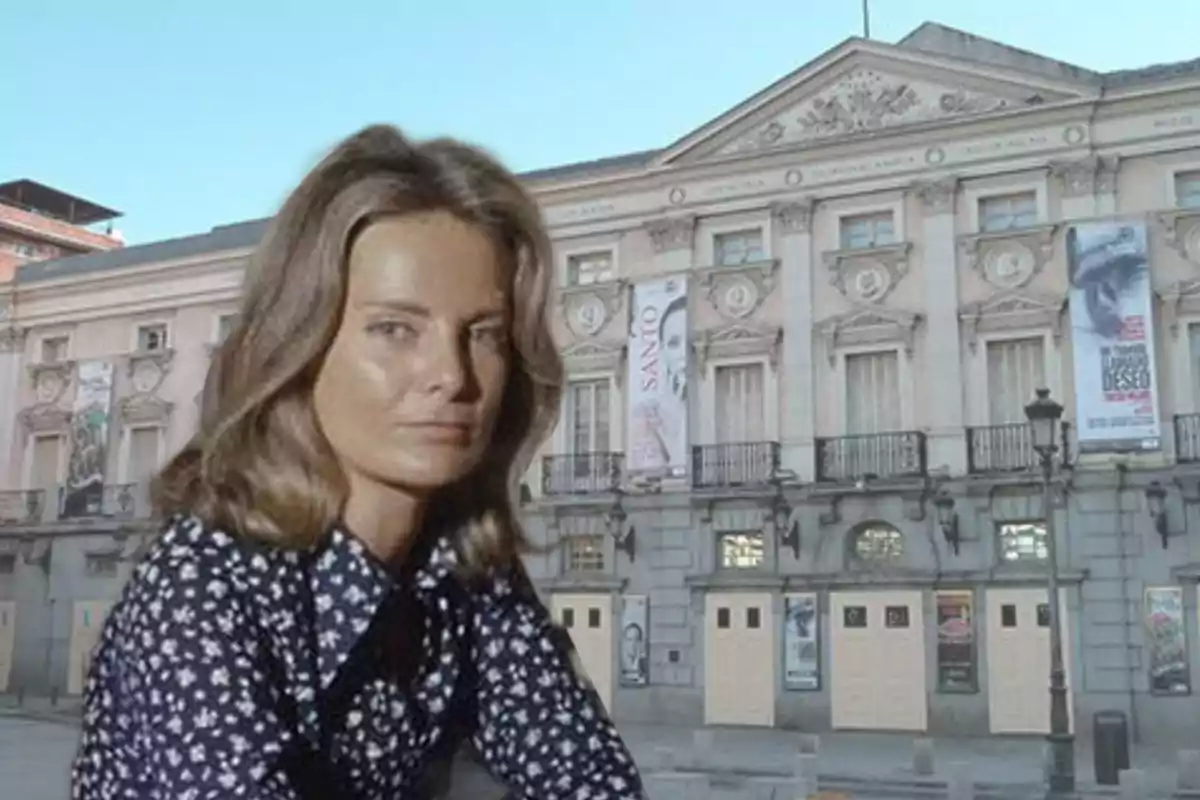 Mujer de cabello castaño con blusa de flores frente a un edificio histórico con carteles en la fachada.