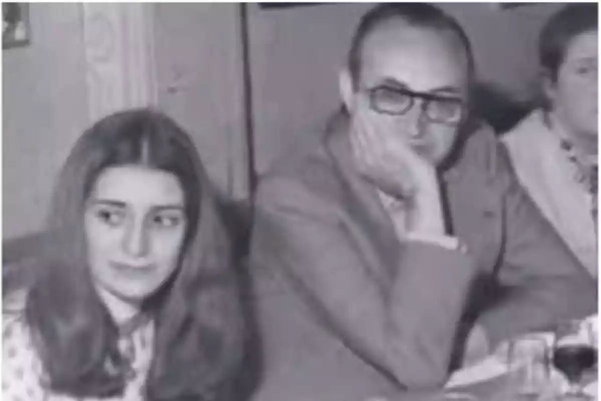 Una imagen en blanco y negro de dos personas sentadas en una mesa, una mujer joven con cabello largo y un hombre con gafas apoyando su cabeza en su mano.