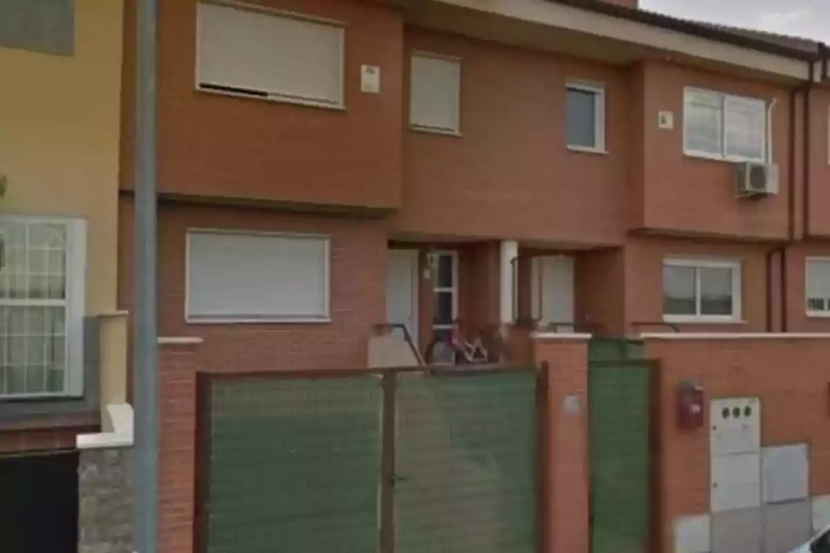 Una casa de ladrillo con ventanas cerradas y una bicicleta en la entrada, rodeada por una cerca verde.