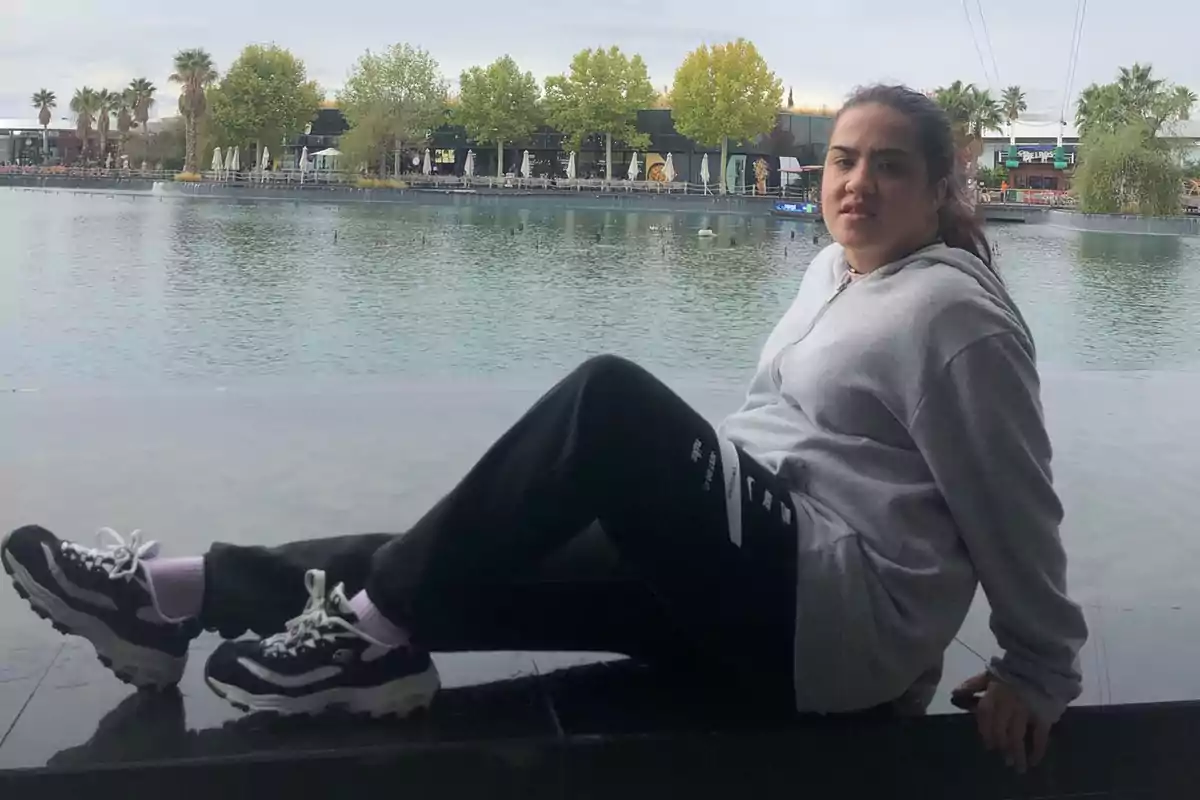 Una persona sentada junto a un lago con árboles y edificios al fondo.