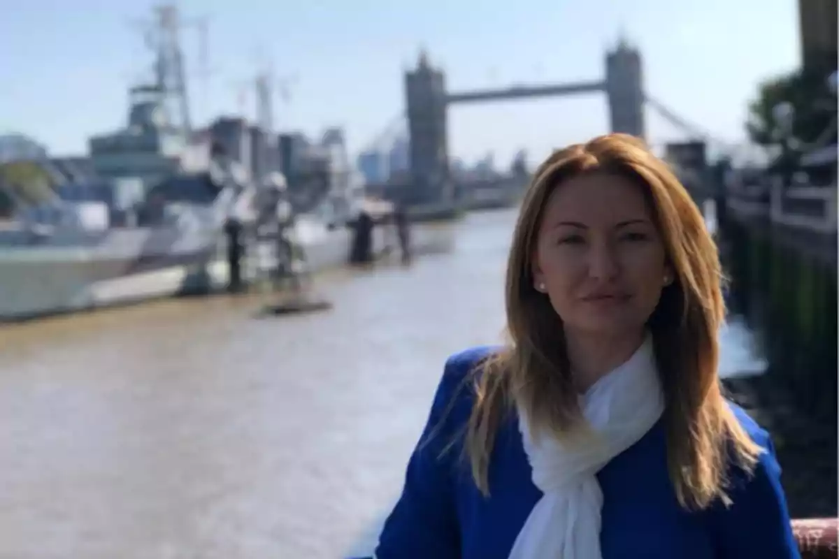 Una mujer con cabello rubio y bufanda blanca posa frente al río Támesis con el Puente de la Torre al fondo.