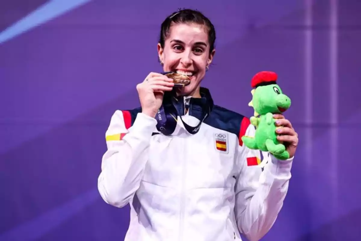 Una deportista con uniforme de España muerde una medalla y sostiene una mascota de peluche verde.