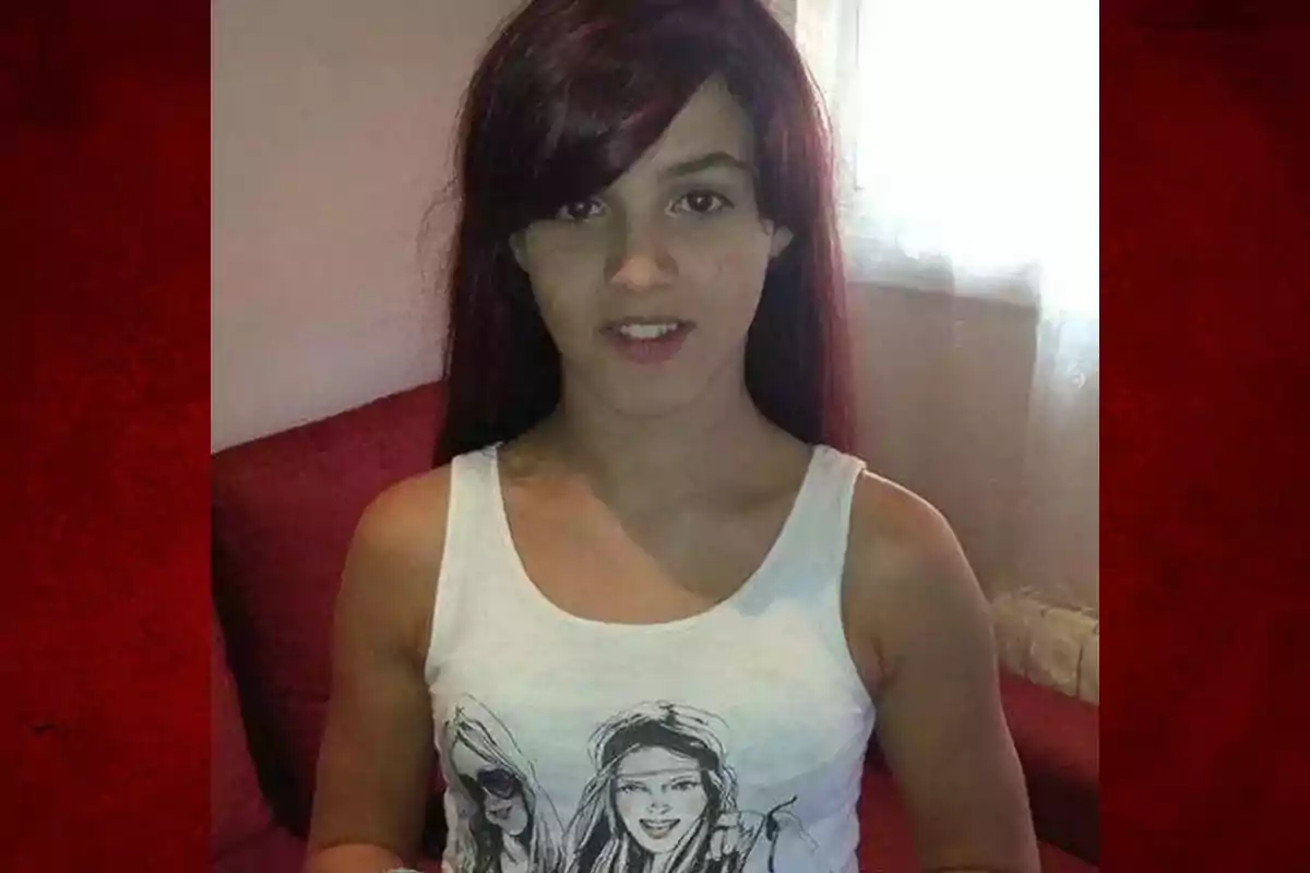 Joven de cabello largo y oscuro con camiseta blanca ilustrada, sentada en un sillón rojo frente a una ventana con cortina blanca.