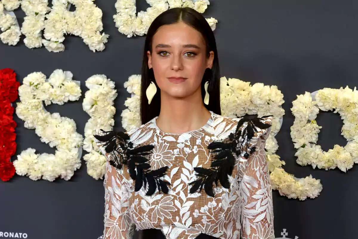 Mujer con vestido de encaje blanco y negro posando frente a un fondo decorado con flores blancas y rojas.