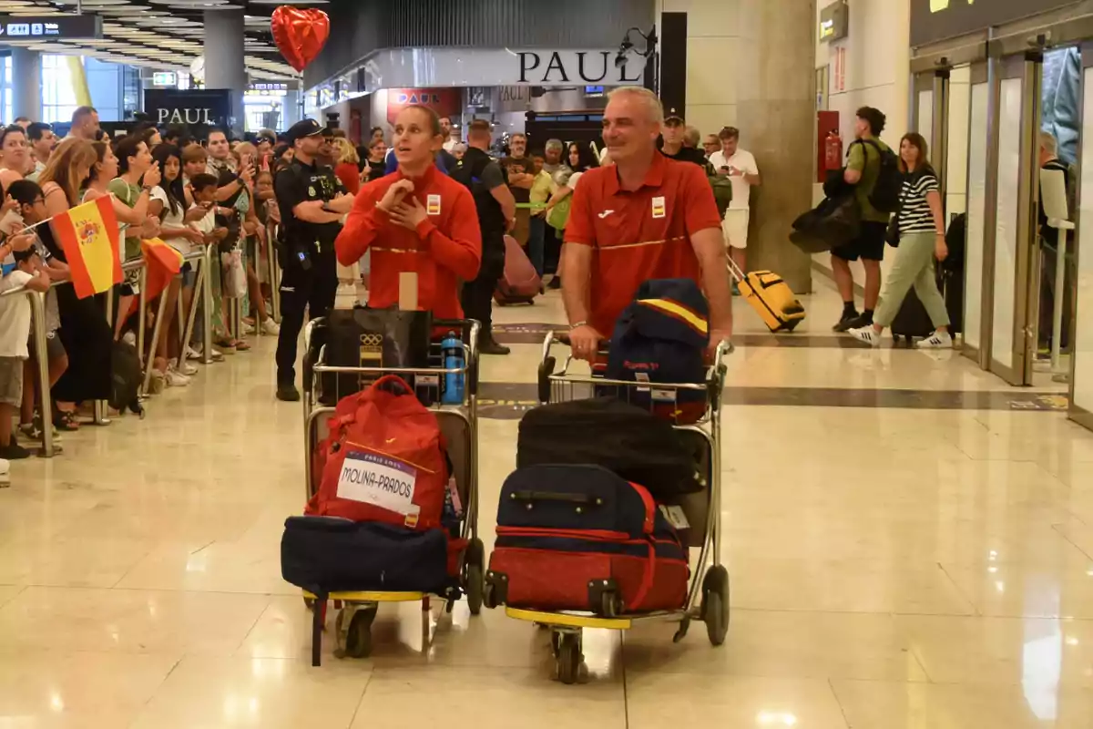 Dos personas con uniformes rojos empujan carritos de equipaje en un aeropuerto, mientras una multitud los recibe con banderas de España y un globo en forma de corazón.