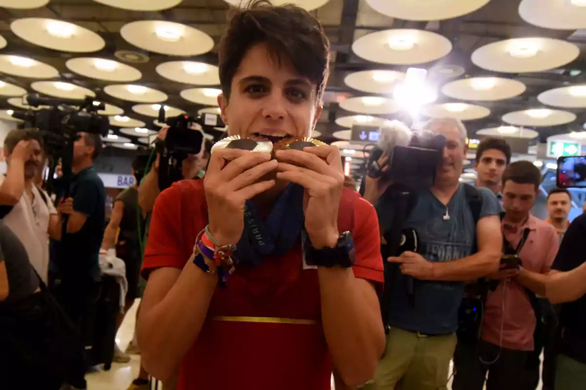 Una persona sostiene dos medallas en la boca mientras es fotografiada por varios reporteros y camarógrafos en un lugar con iluminación de techo circular.