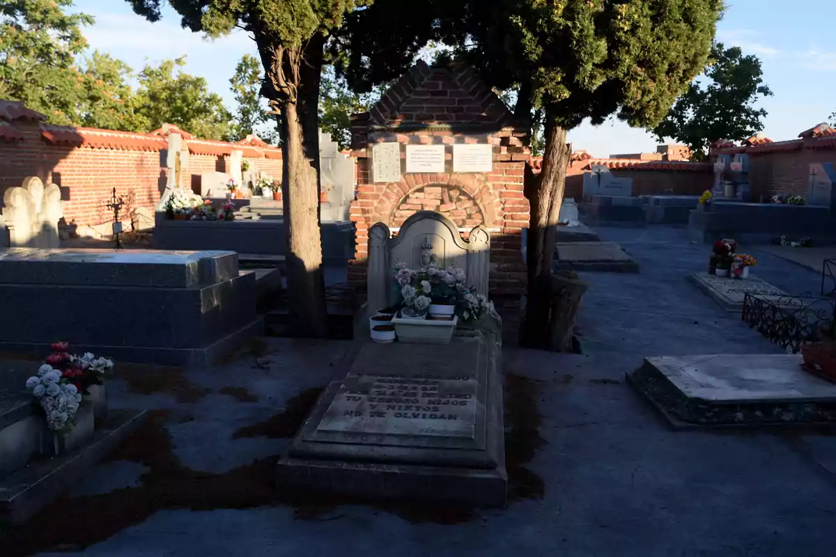 Una tumba en un cementerio con flores y árboles alrededor, con una estructura de ladrillo al fondo.