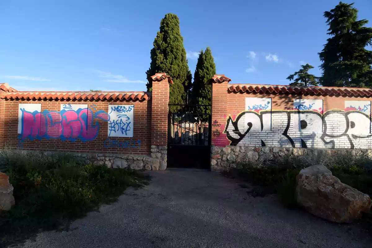 Un muro de ladrillo con grafitis y una puerta de hierro en el centro, rodeado de vegetación y árboles altos bajo un cielo despejado.