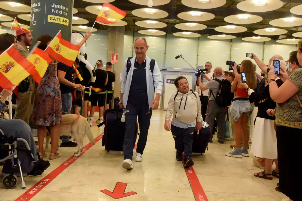 Personas en un aeropuerto reciben a dos hombres con banderas de España y cámaras.