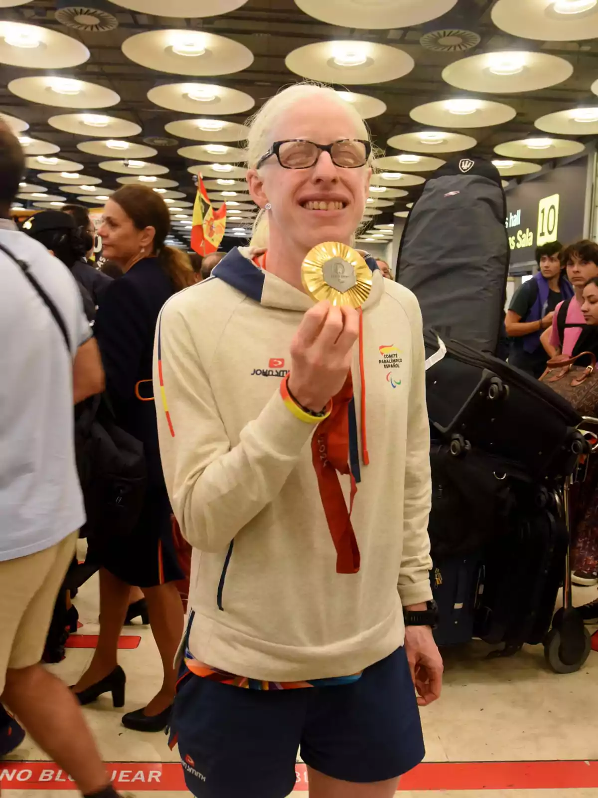 Una persona sonriente sostiene una medalla dorada en un lugar concurrido.