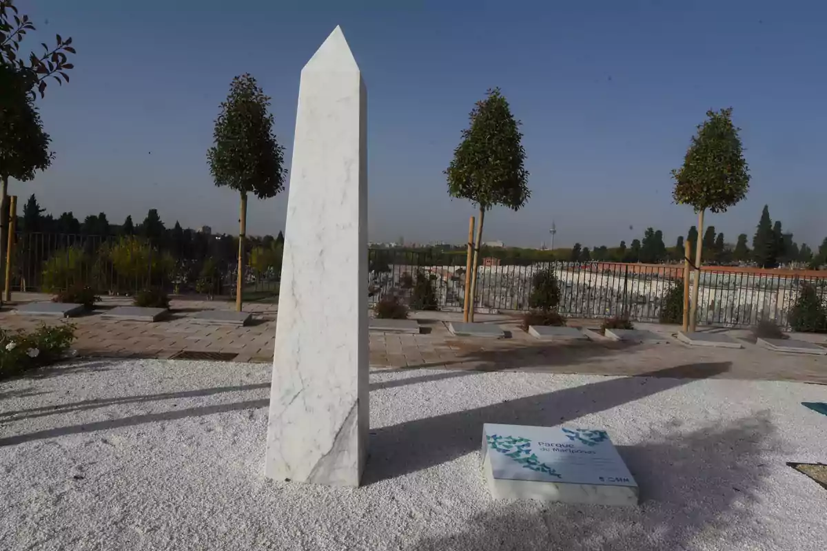 Un obelisco de mármol blanco en un parque rodeado de árboles y un cielo despejado.