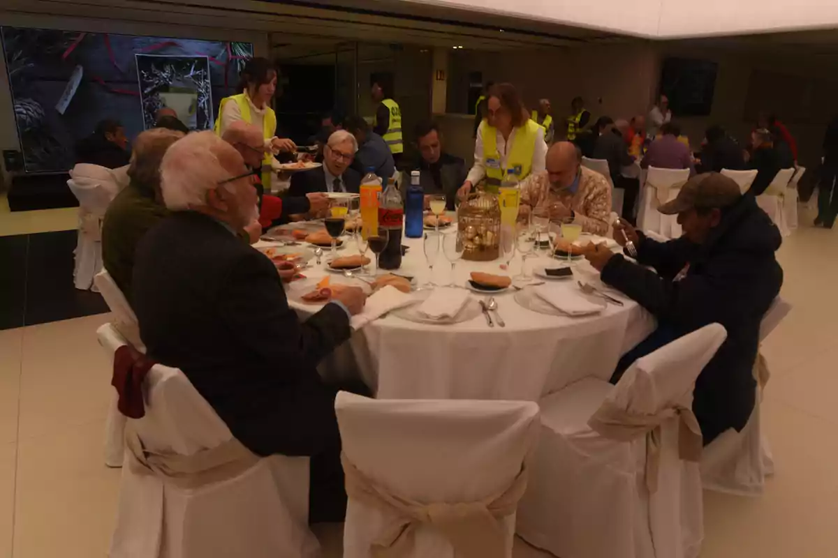 Un grupo de personas mayores comparte una comida en una mesa redonda, atendidos por personal con chalecos amarillos en un ambiente interior.