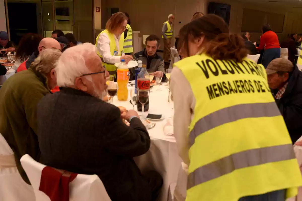 Un grupo de personas se encuentra sentado alrededor de una mesa en un comedor, mientras varios voluntarios con chalecos amarillos los atienden y sirven comida.