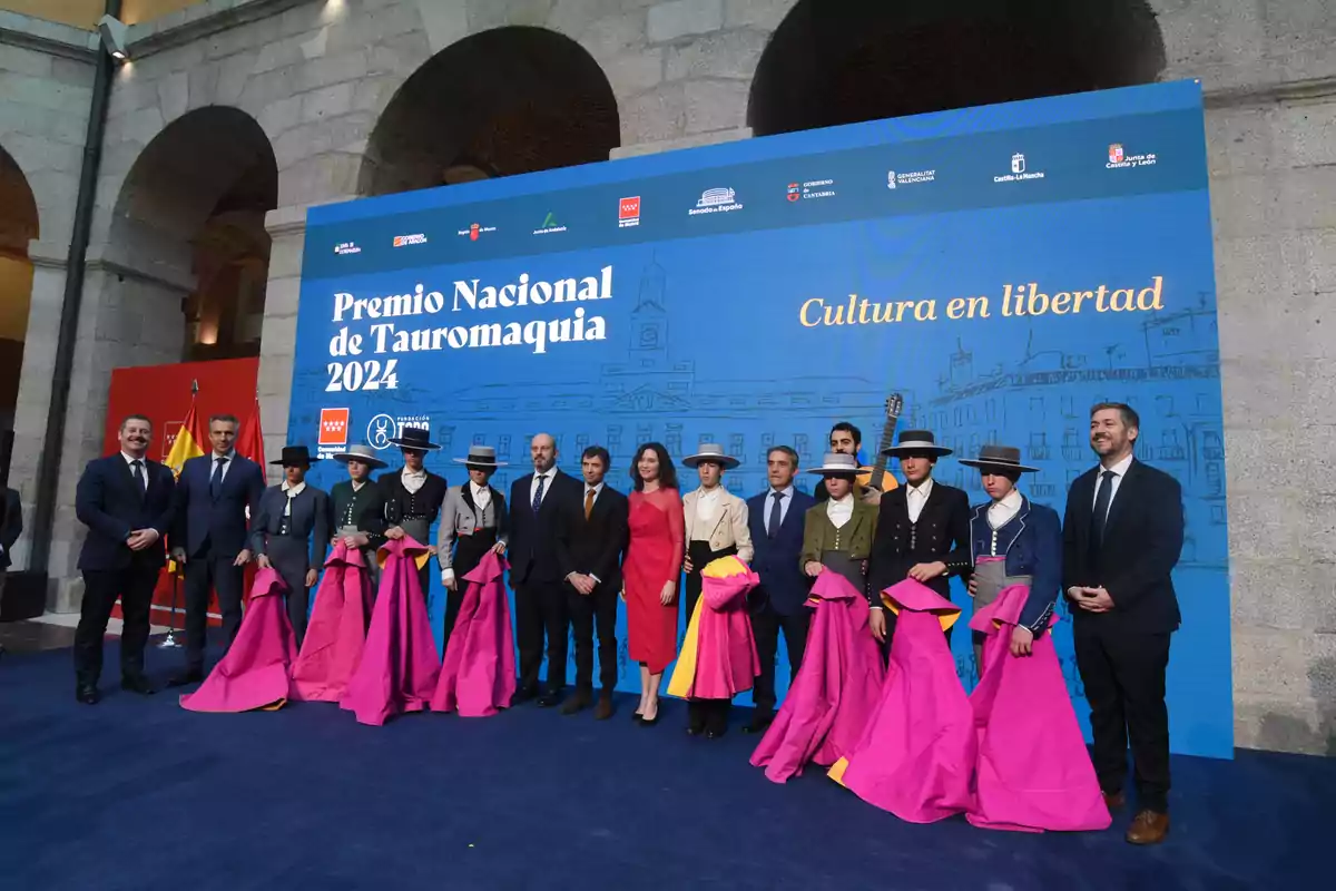 Un grupo de personas posa frente a un cartel del Premio Nacional de Tauromaquia 2024, algunas vestidas con trajes tradicionales y sosteniendo capotes.