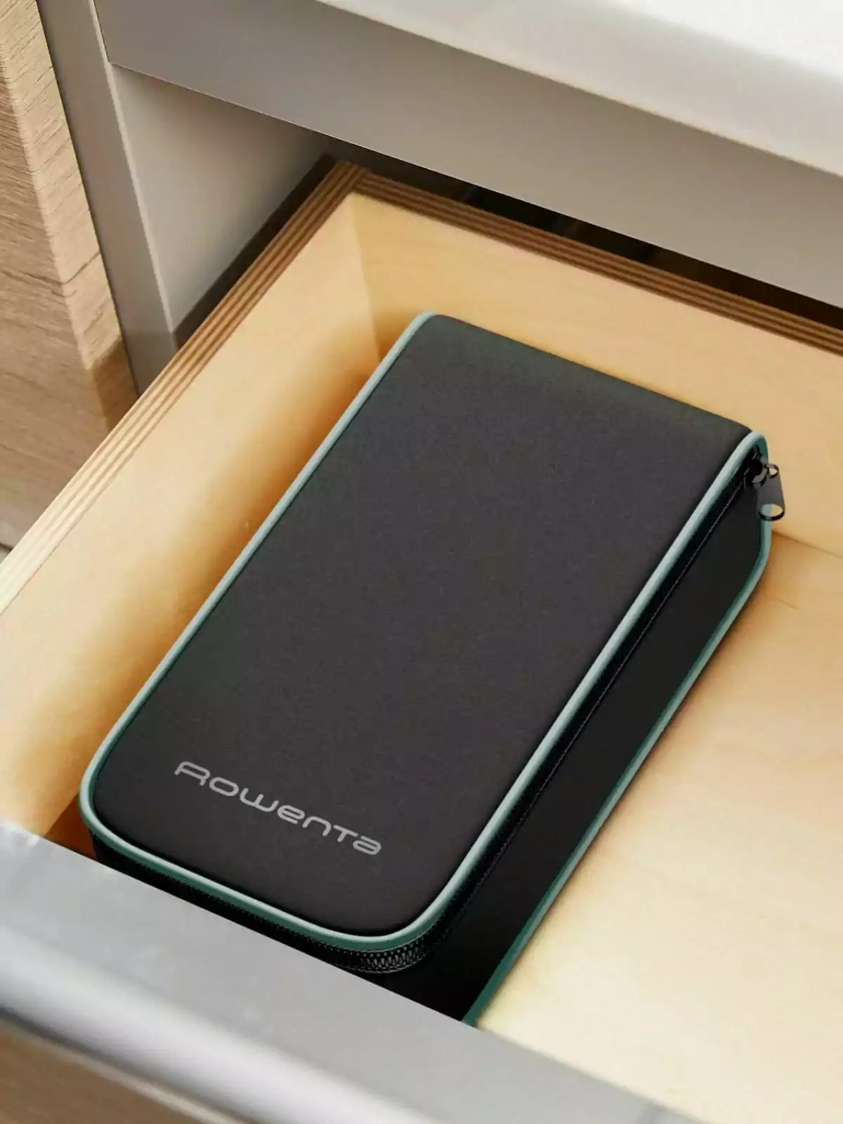 Estuche negro con borde verde y cremallera, con la marca Rowenta, guardado dentro de un cajón de madera clara Estuche negro con borde verde y cremallera, con la marca Rowenta, guardado dentro de un cajón de madera clara