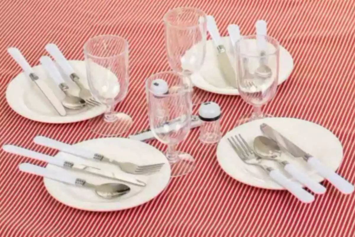 Mesa con mantel rojo a rayas blancas, cuatro platos con cubiertos y copas, salero y pimentero al centro Mesa con mantel rojo a rayas blancas, cuatro platos con cubiertos y copas, salero y pimentero al centro