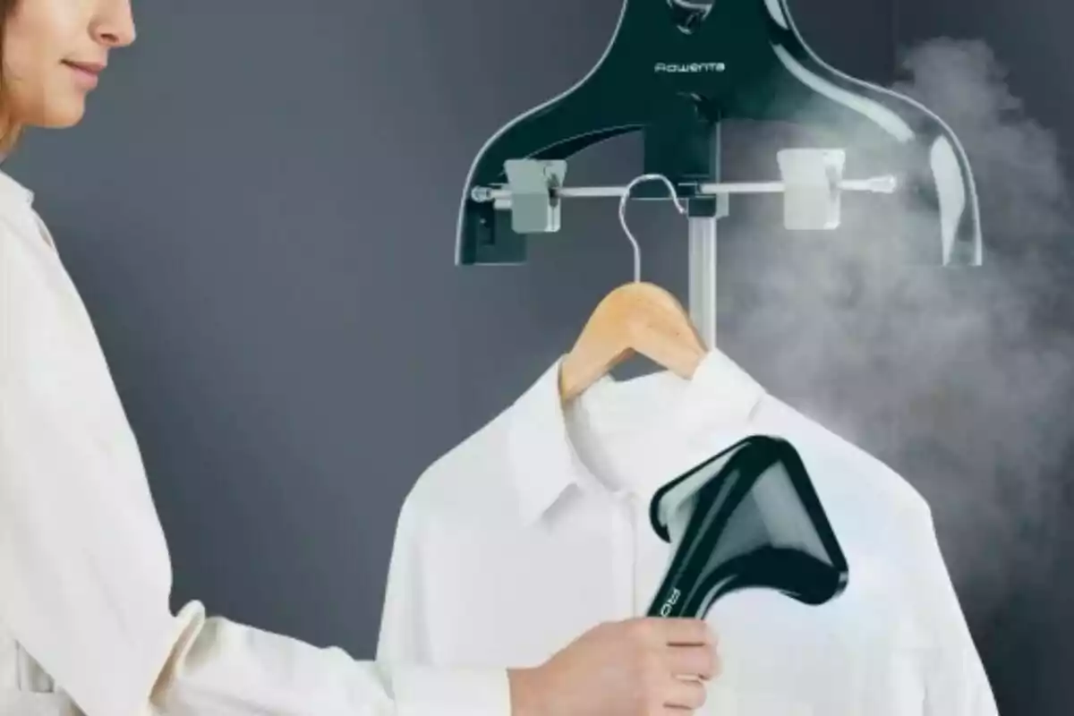 Persona usando un vaporizador de ropa para quitar arrugas de una camisa blanca colgada en una percha Persona usando un vaporizador de ropa para quitar arrugas de una camisa blanca colgada en una percha
