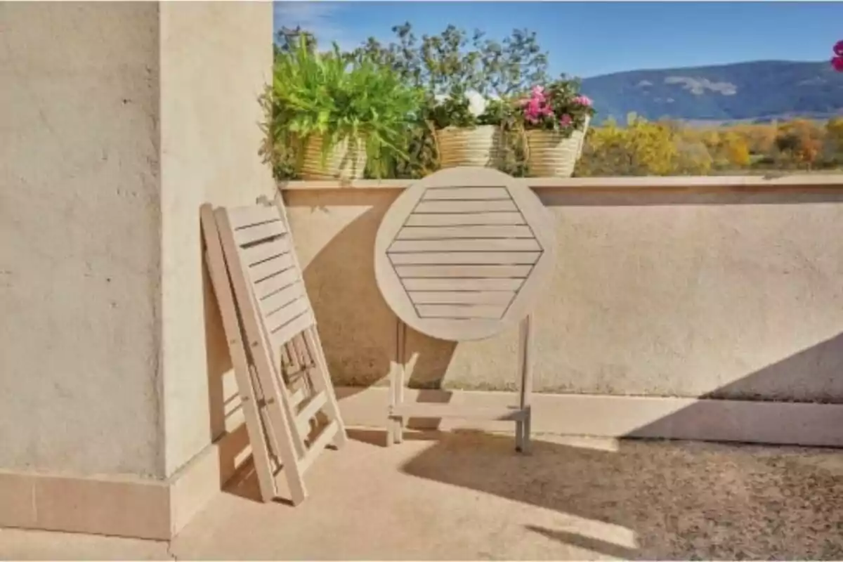 Sillas y mesa plegables de madera apoyadas en una pared de terraza con macetas y vista a las montañas al fondo Sillas y mesa plegables de madera apoyadas en una pared de terraza con macetas y vista a las montañas al fondo