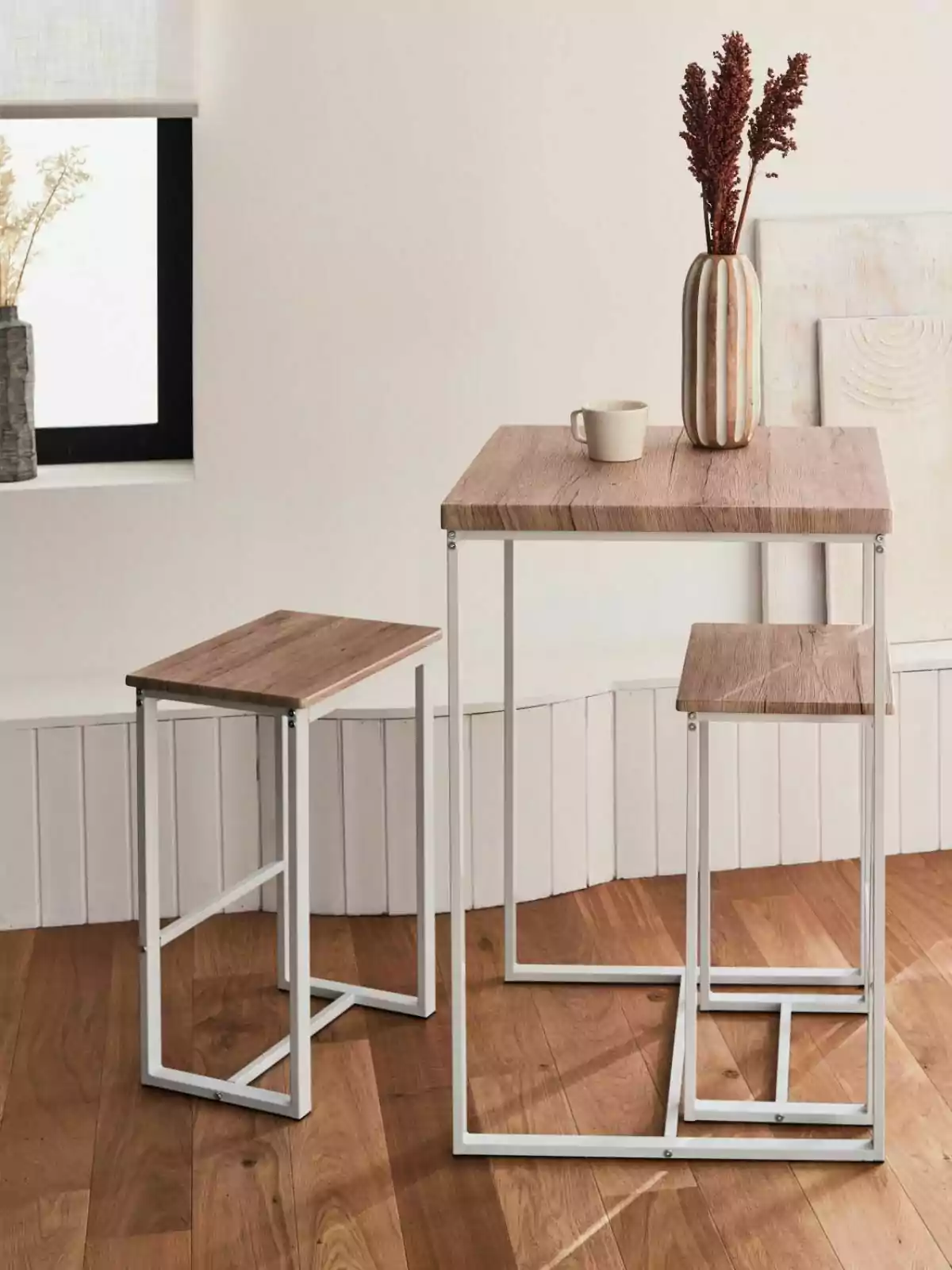 Un conjunto de mesa y taburetes de madera con estructura metálica blanca sobre un suelo de madera, decorado con un jarrón con flores secas y una taza. Un conjunto de mesa y taburetes de madera con estructura metálica blanca sobre un suelo de madera, decorado con un jarrón con flores secas y una taza.