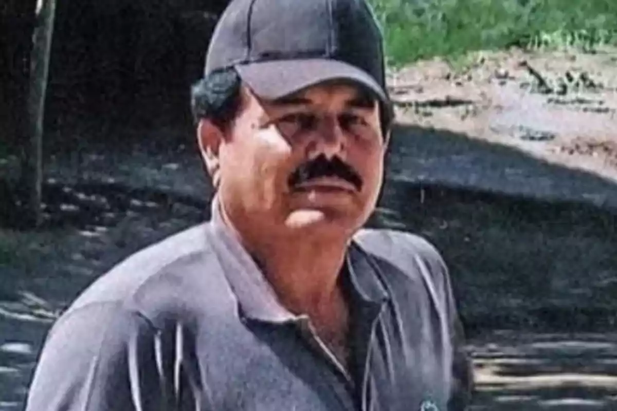 Hombre con bigote y gorra negra, vestido con una camisa gris, en un entorno al aire libre.