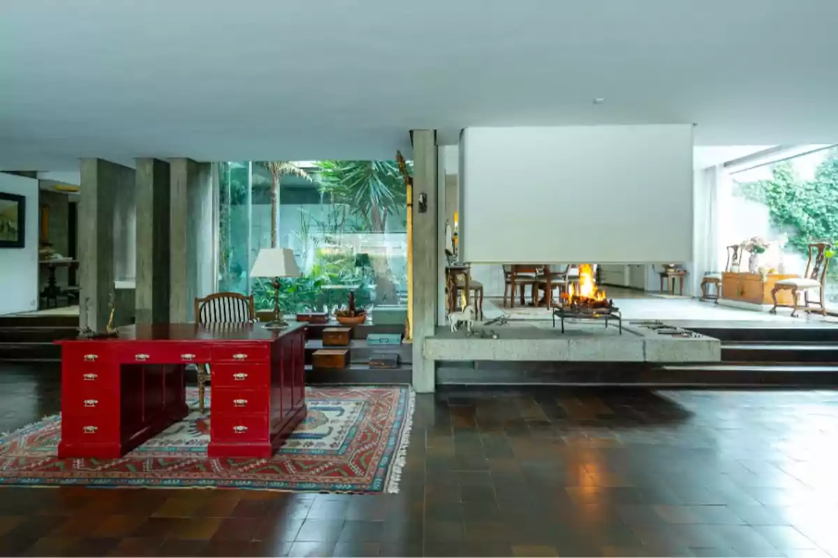 Sala de estar con escritorio rojo, alfombra decorativa, chimenea encendida y grandes ventanales con vista al jardín.