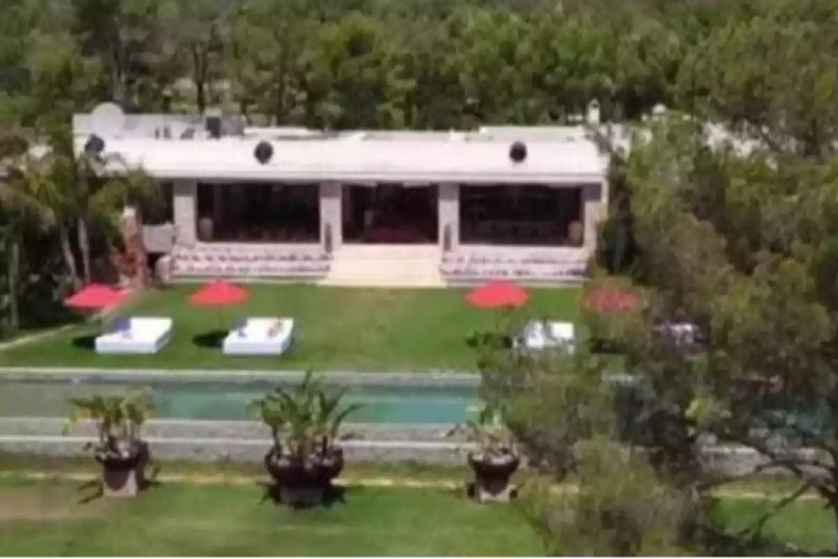 Una casa moderna con un amplio jardín, piscina y sombrillas rojas rodeada de árboles.