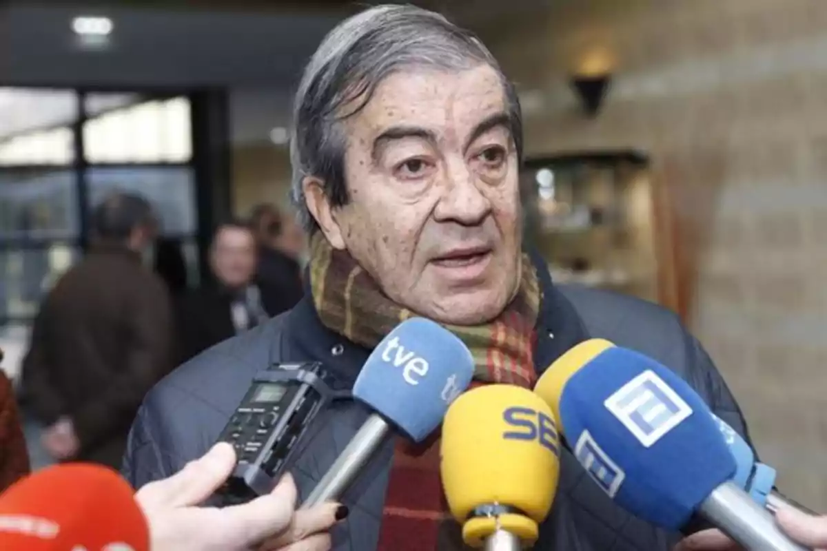 Hombre mayor con bufanda siendo entrevistado por varios medios de comunicación con micrófonos de colores frente a él