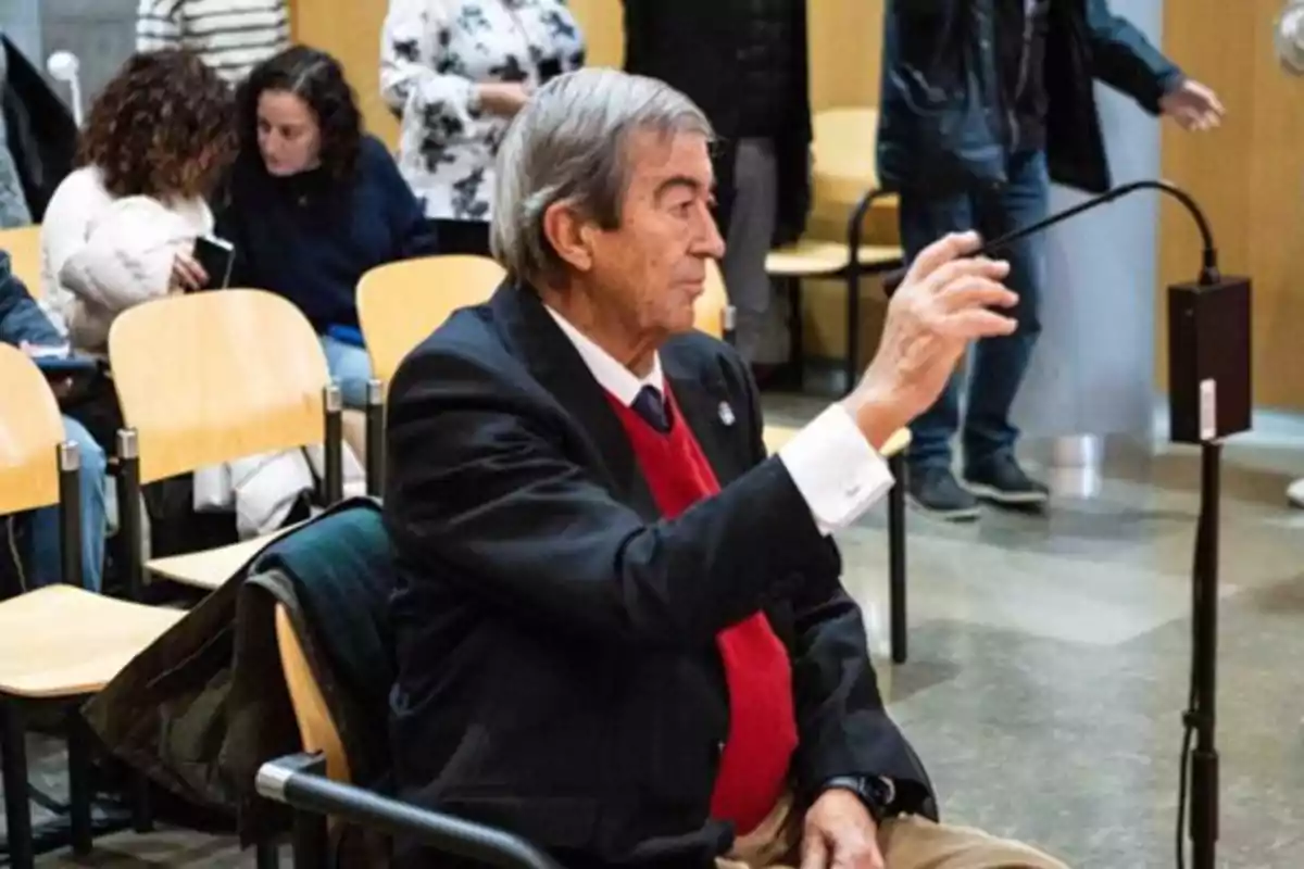 Hombre mayor de cabello canoso y suéter rojo sentado en una silla dentro de una sala con otras personas al fondo Hombre mayor de cabello canoso y suéter rojo sentado en una silla dentro de una sala con otras personas al fondo