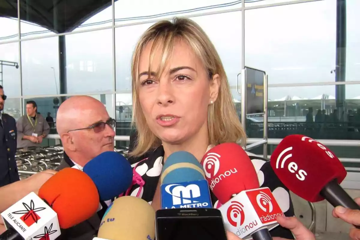 Una mujer hablando frente a varios micrófonos de diferentes medios de comunicación en un entorno exterior. Una mujer hablando frente a varios micrófonos de diferentes medios de comunicación en un entorno exterior.