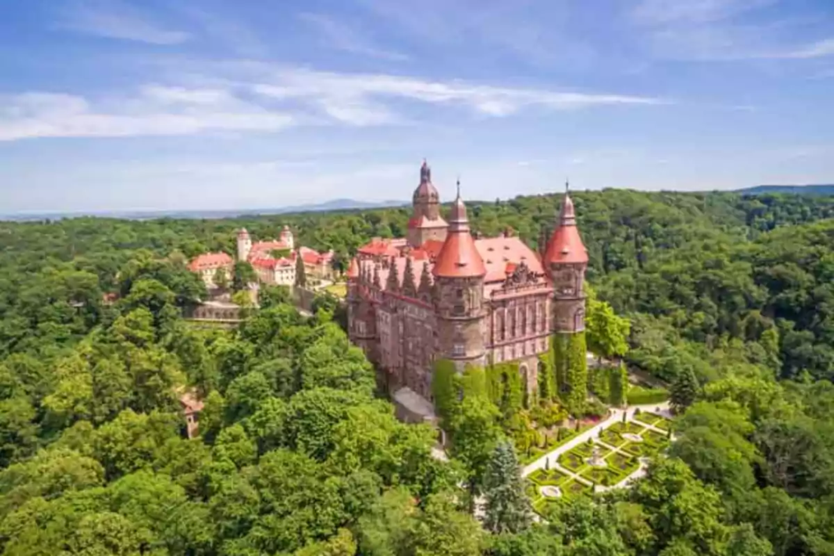 Vista aérea de un castillo rodeado de un frondoso bosque y jardines bien cuidados bajo un cielo azul.