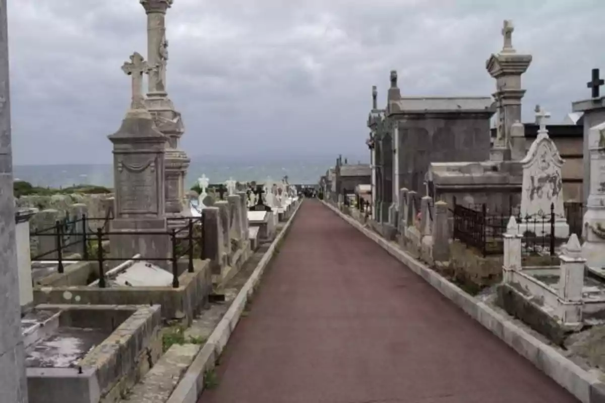 Un camino pavimentado atraviesa un cementerio con tumbas y mausoleos de piedra a ambos lados bajo un cielo nublado.