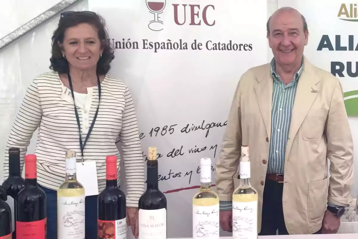 Dos personas sonrientes posan detrás de una mesa con varias botellas de vino en un evento de la Unión Española de Catadores Dos personas sonrientes posan detrás de una mesa con varias botellas de vino en un evento de la Unión Española de Catadores