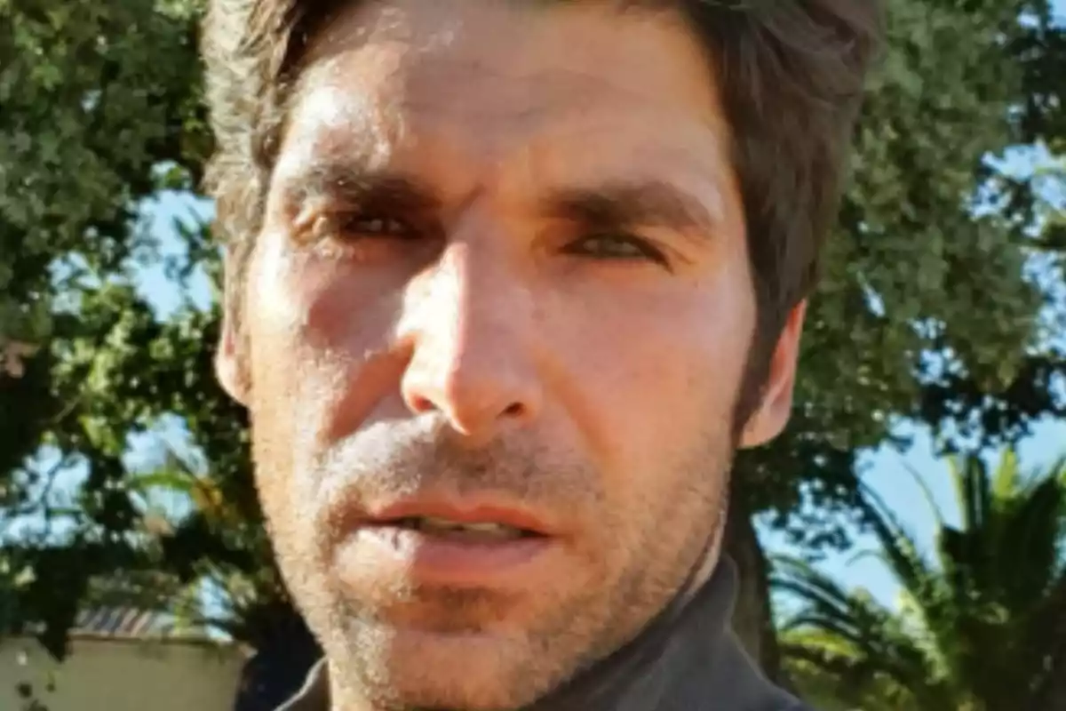 Un hombre con barba ligera y cabello oscuro en un entorno al aire libre con árboles al fondo.